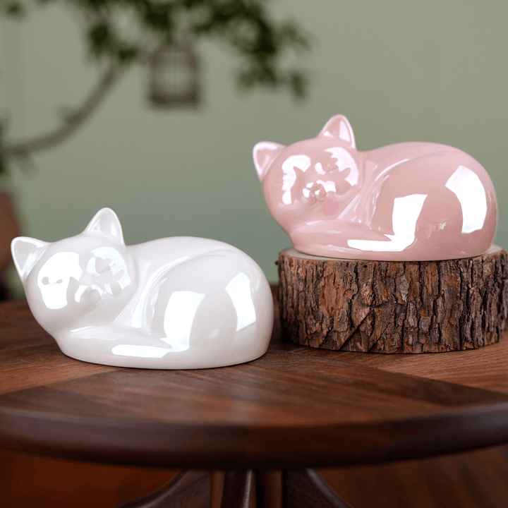 Two ceramic cat figurines, one white and one pink, on a wooden surface with a blurred green background.